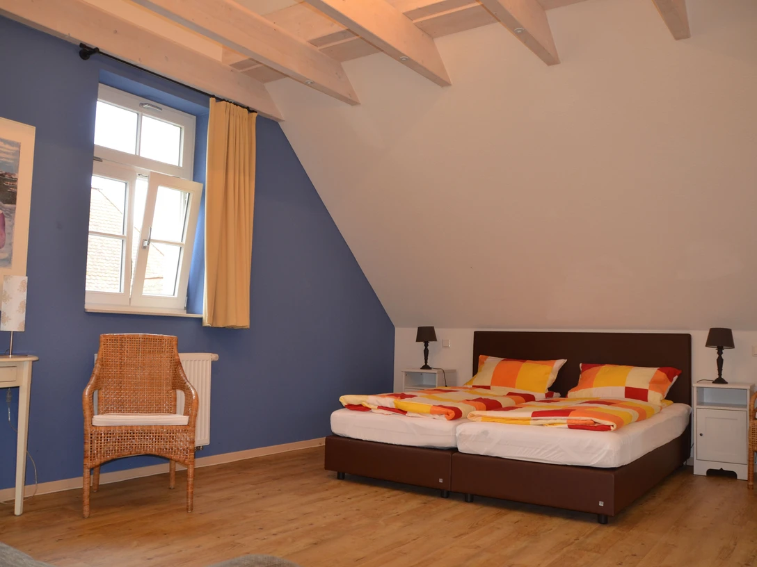 Ein gemütliches Doppelzimmer mit blauen Wänden, Holzdecke, einem Fenster und orange-gelben Bettdecken.