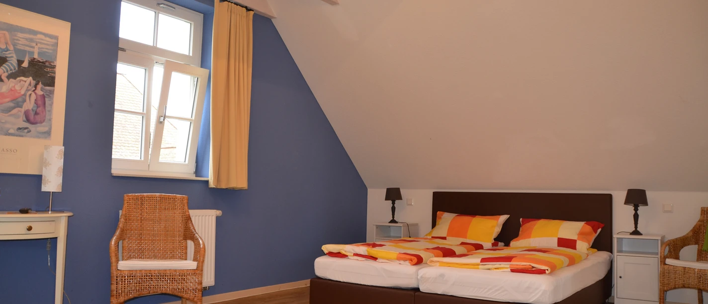 Ein gemütliches Doppelzimmer mit blauen Wänden, Holzdecke, einem Fenster und orange-gelben Bettdecken.