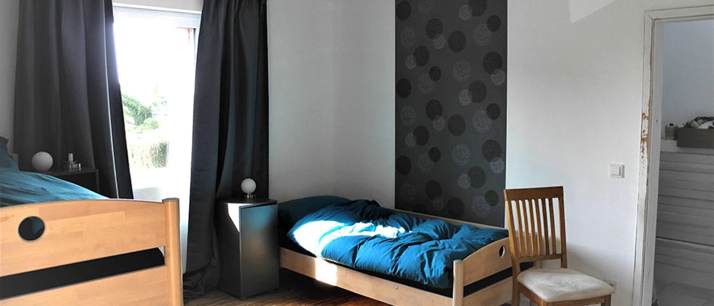 Helles Zimmer mit zwei Holzbetten, einem Stuhl und einem Fenster, das Licht auf den Holzboden wirft.