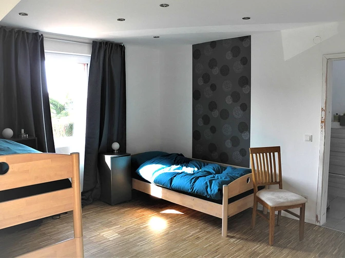 Helles Zimmer mit zwei Holzbetten, einem Stuhl und einem Fenster, das Licht auf den Holzboden wirft.