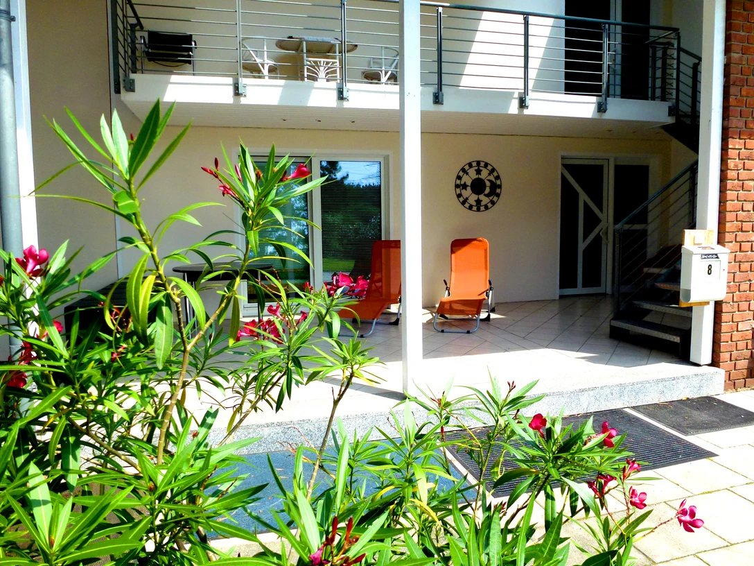 Terrasse einer Ferienwohnung mit Sonnenliegen, umgeben von blühenden Pflanzen und modernem Mobiliar.