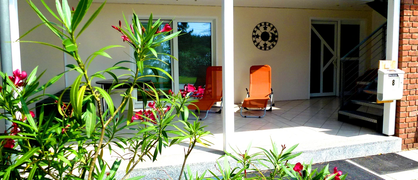 Terrasse einer Ferienwohnung mit Sonnenliegen, umgeben von blühenden Pflanzen und modernem Mobiliar.