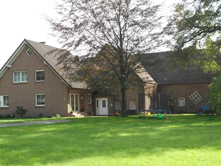 FeWo Stallmanns Hof.jpg Backsteinhaus mit Satteldach auf grünem Rasen, umgeben von Bäumen, Vorderseite mit Eingangstür