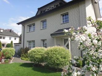 FeWo Ritter20130511_0021.jpg Ein zweistöckiges Haus mit grauem Walmdach und heller Klinkerfassade, umgeben von blühendem Garten.