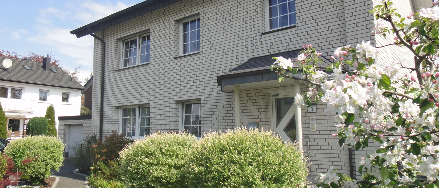 FeWo Ritter20130511_0021.jpg Ein zweistöckiges Haus mit grauem Walmdach und heller Klinkerfassade, umgeben von blühendem Garten.