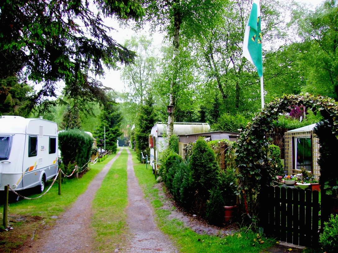 Ein Campingplatz mit Wohnwagen und gepflegten Hecken, flankiert von einem begrünten Weg und einer Fahne.