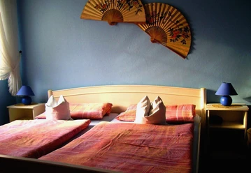 Pension_Sennetreff__StadtSHS.JPG Ein Doppelbett mit rosa Bettwäsche, flankiert von blauen Lampen und asiatischen Fächern an der Wand.