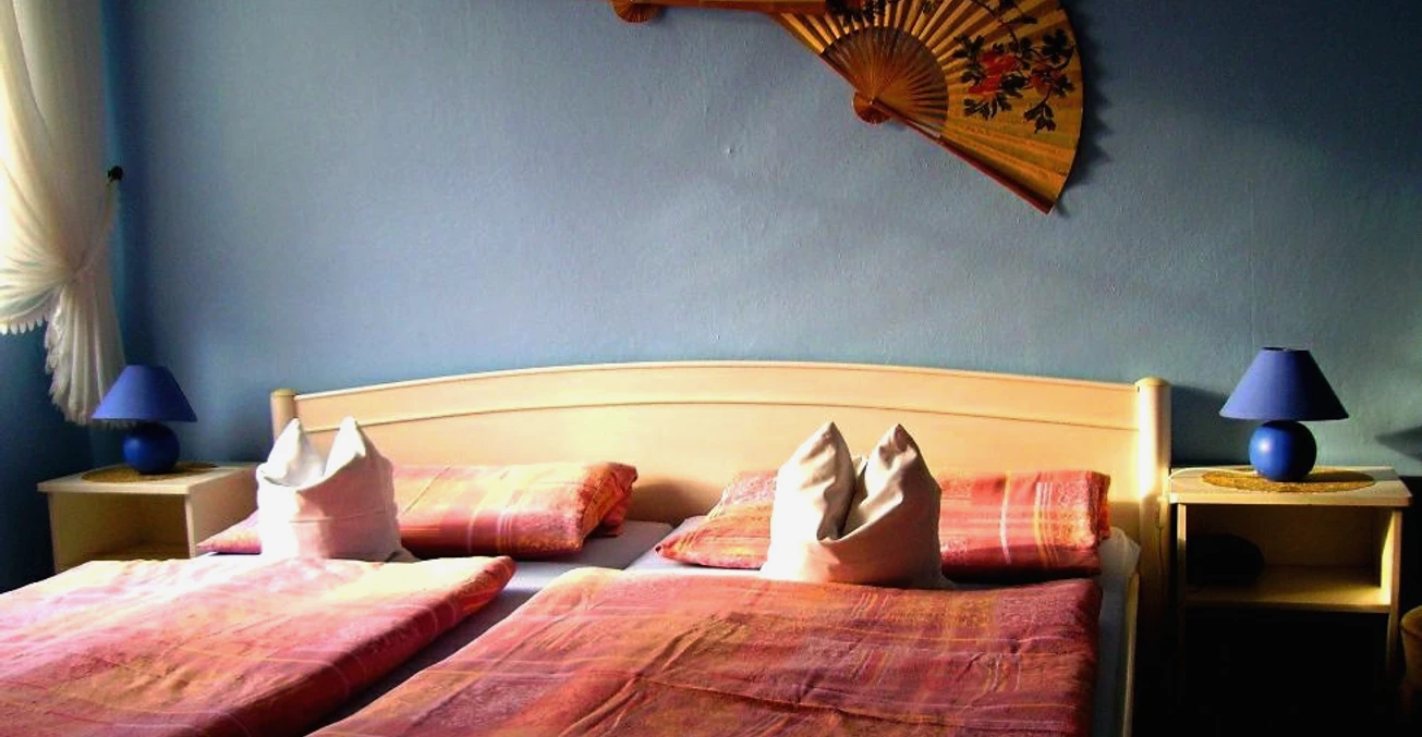 Pension_Sennetreff__StadtSHS.JPG Ein Doppelbett mit rosa Bettwäsche, flankiert von blauen Lampen und asiatischen Fächern an der Wand.