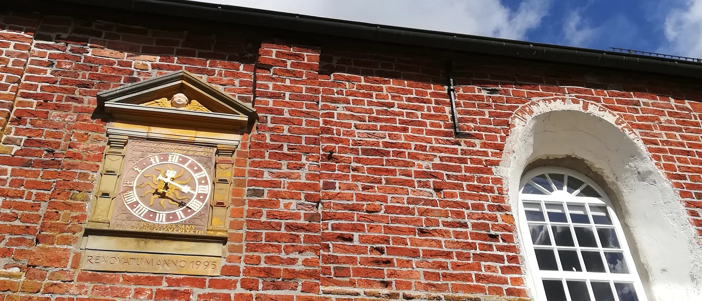 Kirche Freepsum Rote Backsteinfassade der Kirche Freepsum mit Turmuhr und großem Fenster unter blauem Himmel.