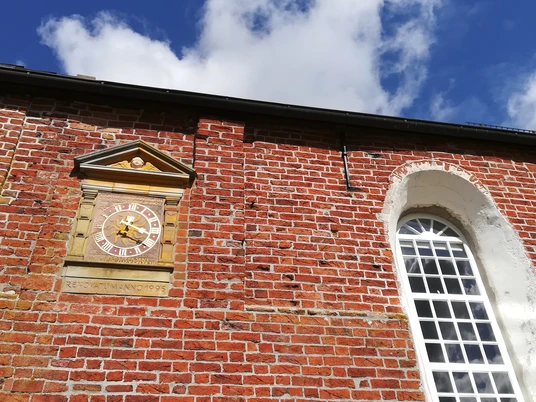 Kirche Freepsum Rote Backsteinfassade der Kirche Freepsum mit Turmuhr und großem Fenster unter blauem Himmel.