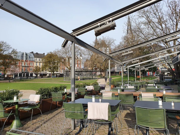 Von Blumenkübeln begrenzte Außenterrasse mit vielen Tischen und Stühlen mit Decken und Blick auf den Elisengarten.