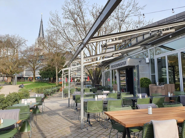 Terasse 2 (c) Restaurant Elisenbrunnen.jpg Von Blumenkübeln begrenzte Außenterrasse mit vielen Tischen und Stühlen mit Decken und Blick auf den Eingang des Restaurants und den Elisengarten.