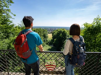 Halle-HagedornDenkmal-Teutoburger-Wald-Tourismus-Patrick-Gawandtka-093_lowres.jpg Zwei Wanderer mit Rucksäcken blicken von einer Balustrade auf eine weite, grüne Landschaft.