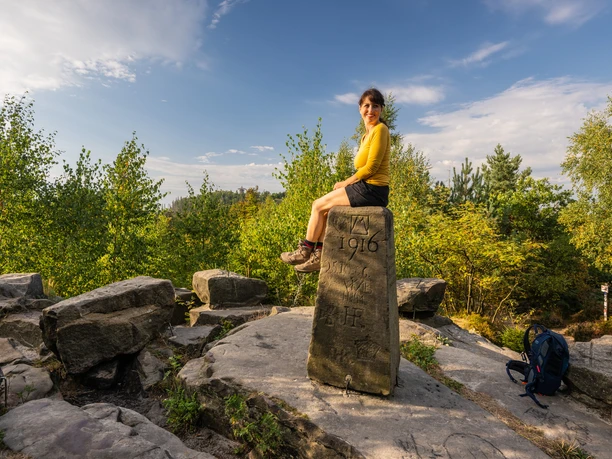 Frau sitzt auf historischem Grenzstein umgeben von Bäumen und bewaldeter Landschaft unter blauem Himmel.