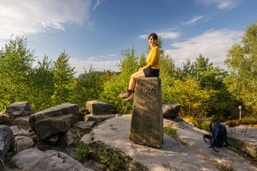 Lippischer Velmerstot-Kreis Lippe-Teutoburger-Wald-Tourismus-D-Ketz-001.jpg Frau sitzt auf historischem Grenzstein umgeben von Bäumen und bewaldeter Landschaft unter blauem Himmel.