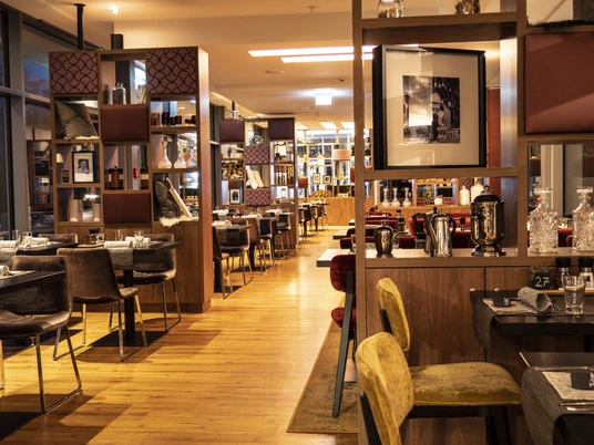 Leander Bad Oeynhausen Zu sehen ist das Innere des stillvollen Restaurants Leander mit warmen und gemütlichen Licht. Die Einrichtung besteht aus dekorativen Regalen, elegant gedeckte tischen und Wandelelementen mit Bildern, was eine einladende Atmosphäre schafft.