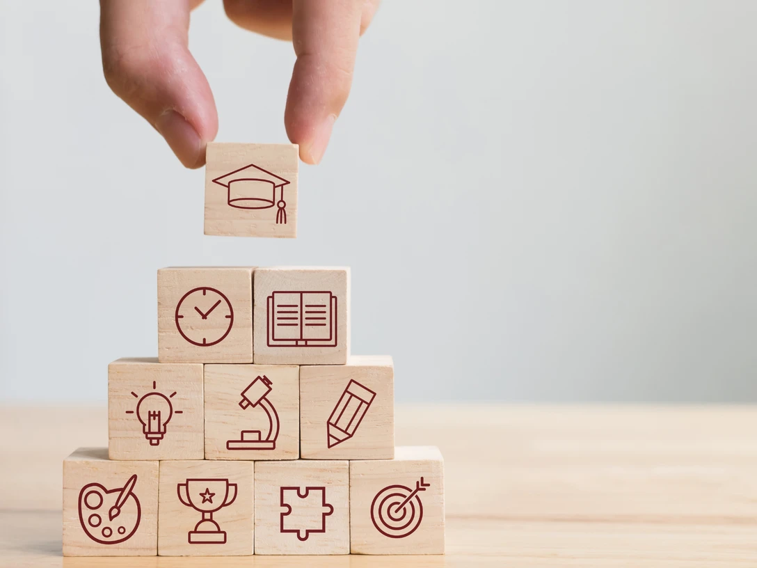 Stockbild VHS Eine Hand platziert einen Holzblock mit Hut-Icon auf Würfel, die Bildungssymbole zeigen.