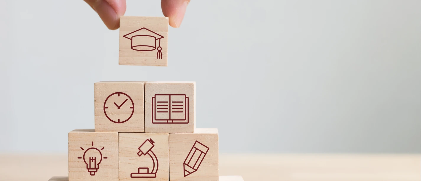 Stockbild VHS Eine Hand platziert einen Holzblock mit Hut-Icon auf Würfel, die Bildungssymbole zeigen.