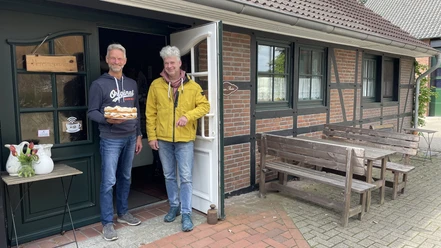 Eingangsbereich Hof Cafe Janßen Zwei freundliche Männer stehen vor einem Backsteinhaus; einer hält eine Torte, lächelnd in die Kamera.