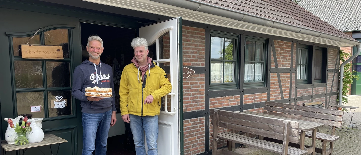 Eingangsbereich Hof Cafe Janßen Zwei freundliche Männer stehen vor einem Backsteinhaus; einer hält eine Torte, lächelnd in die Kamera.