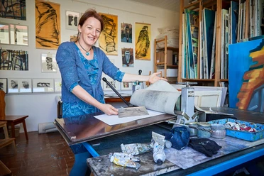 Künstlerin Anne Kern Eine Frau in einem blauen Kleid steht in einem Atelier vor einer Druckpresse, umgeben von Kunstwerken und Malutensilien.