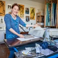 Künstlerin Anne Kern Eine Frau in einem blauen Kleid steht in einem Atelier vor einer Druckpresse, umgeben von Kunstwerken und Malutensilien.