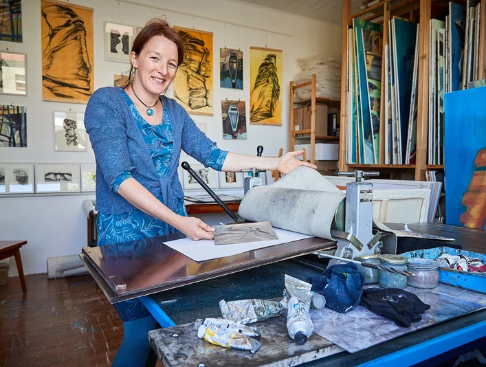 Künstlerin Anne Kern Eine Frau in einem blauen Kleid steht in einem Atelier vor einer Druckpresse, umgeben von Kunstwerken und Malutensilien.
