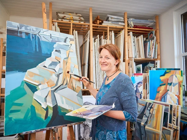 Anne Kern beim Malen Eine Frau malt in einem Atelier ein abstraktes Bild in Blau- und Beigetönen. Sie lächelt, umgeben von weiteren Gemälden und Leinwänden.