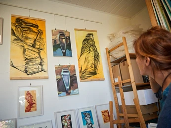 Anne Kern - Kunstmeile Wehlen Eine Frau betrachtet in einem Atelier mehrere abstrakte Gemälde in Gelb- und Blautönen, die an einer weißen Wand hängen.