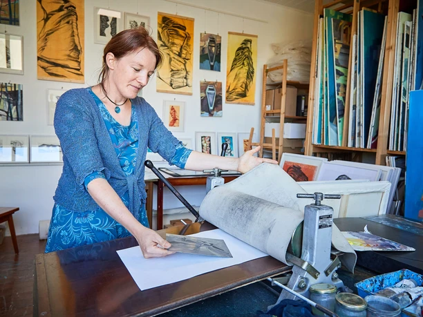 Künstlerin Anne Kern Eine Frau in blauer Kleidung arbeitet in einem Atelier an einer Druckpresse, umgeben von Kunstwerken und Malutensilien.