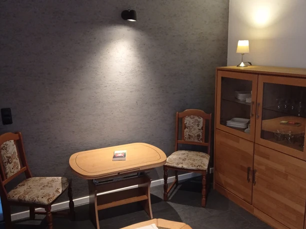 IMG_20230118_155127.jpg Gemütlicher Innenraum mit zwei Holzstühlen, kleinem Tisch, Schrank und dezentem Beleuchtungs-Setup.