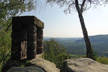 Bekijk Hohe Liebe Stenen uitkijkpunt met uitzicht op beboste heuvels en rotsen op de achtergrond, omringd door bomen onder een heldere hemel.