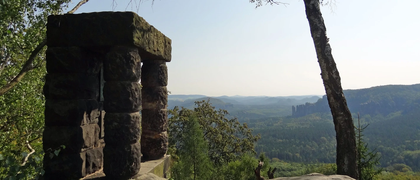 View Hohe Liebe Stone viewpoint with a view of wooded hills and rocks in the background, surrounded by trees under a clear sky.