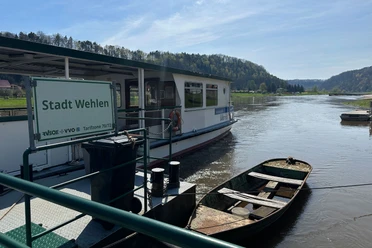Fähre Wehlen Fähre "Stadt Wehlen" auf einem Fluss, umgeben von grünen Hügeln und blauem Himmel, mit einem kleinen Boot im Vordergrund.