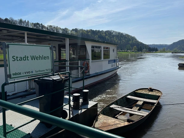 Fähre Wehlen Fähre "Stadt Wehlen" auf einem Fluss, umgeben von grünen Hügeln und blauem Himmel, mit einem kleinen Boot im Vordergrund.
