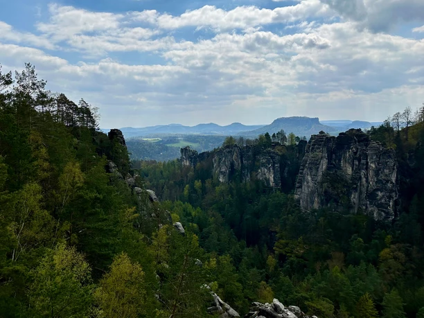 View of a wooded rocky landscape with steep cliffs under a cloudy sky, with rolling hills in the background.