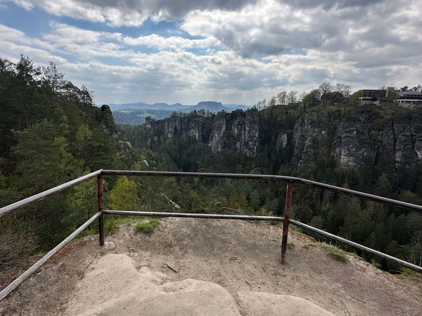 Viewpoint with rusty railings, surrounded by green trees and steep rocks under a cloudy sky, mountains in the background.