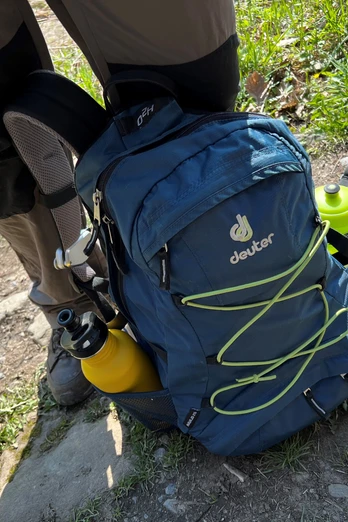 Outdoor-Rucksack in Blau mit gelben Flaschen an den Seiten auf einem Wanderweg zwischen Gras und Steinen.