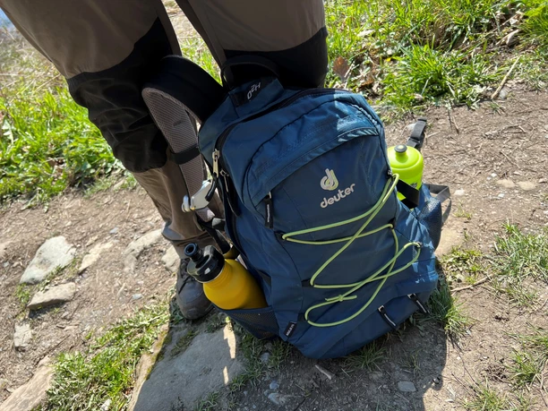 Outdoor backpack in blue with yellow bottles on the sides on a hiking trail between grass and stones.