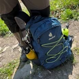 Rucksack Outdoor-Rucksack in Blau mit gelben Flaschen an den Seiten auf einem Wanderweg zwischen Gras und Steinen.