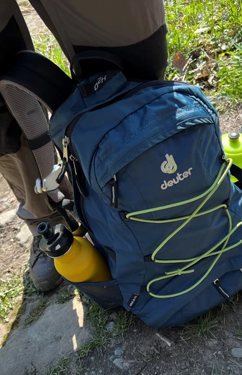 Rucksack Outdoor-Rucksack in Blau mit gelben Flaschen an den Seiten auf einem Wanderweg zwischen Gras und Steinen.