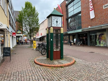Spargeluhr Eine markante Turmuhr aus Metall mit grünen und goldenen Akzenten steht in einer belebten Fußgängerzone.