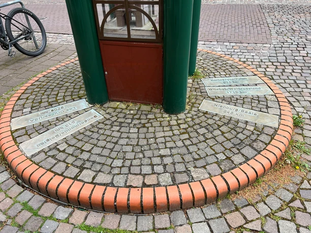Runde Skulptur mit Uhr aus Metall auf einem gepflasterten Platz, umgeben von mehreren Gedenkplatten.