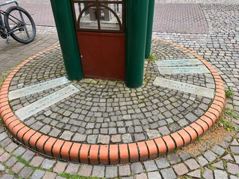 Spargeluhr Runde Skulptur mit Uhr aus Metall auf einem gepflasterten Platz, umgeben von mehreren Gedenkplatten.