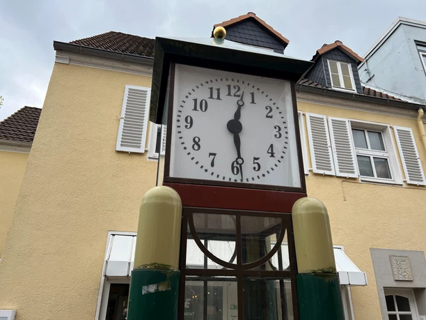 Eine antike Standuhr mit einem großen Ziffernblatt steht vor einem pastellgelben Gebäude mit weißen Fensterläden.