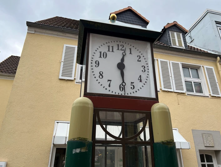 Spargeluhr Eine antike Standuhr mit einem großen Ziffernblatt steht vor einem pastellgelben Gebäude mit weißen Fensterläden.