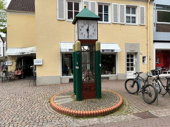 Spargeluhr Eine in ein Uhrwerk integrierte Kunststoffspargel-Statue mit umgebenden Geschäften und Fahrrädern.