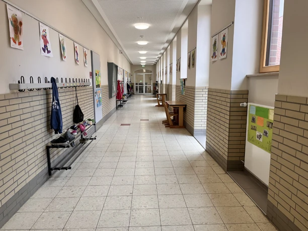IMG_0759.jpg Heller Schulflur mit Garderobenleisten, bunten Kinderzeichnungen an den Wänden und einem Fliesboden.