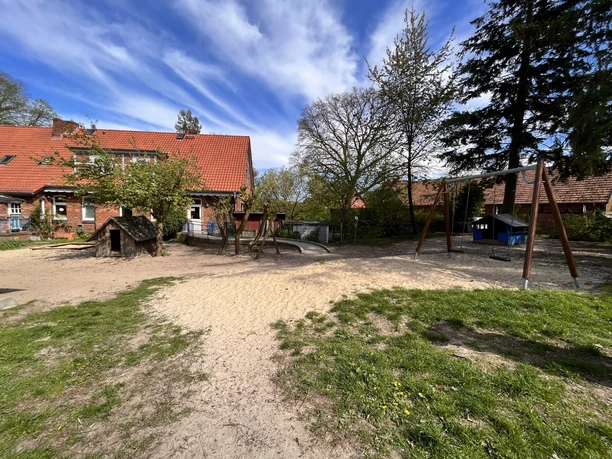 IMG_0770.jpg Außenbereich des Kindergartens mit Sandspielplatz, Schaukel, Holzhaus und Backsteingebäude.