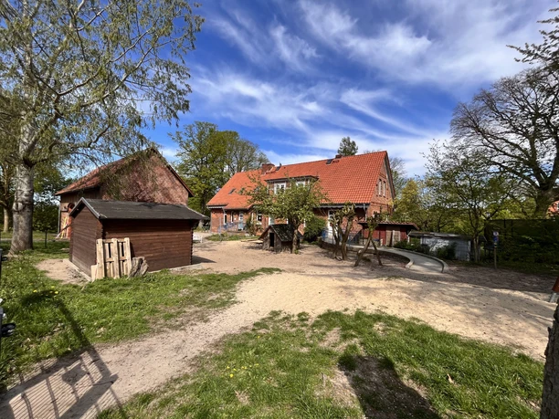 IMG_0771.jpg Klassisches Backsteingebäude mit rotem Ziegeldach und Sandspielplatz im Vordergrund.